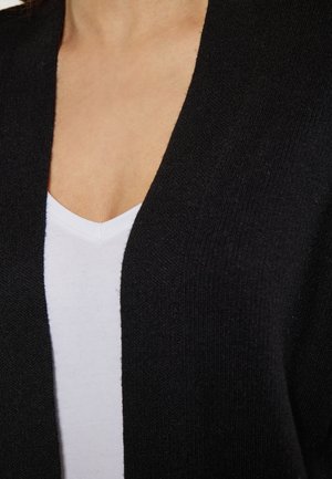 Musta neuletakki, jossa on pehmeä rakenne ja avonainen etuosa, yhdistetty valkoiseen V-kaula-aukkopaitaan, mikä tuo esiin kontrastivärit.