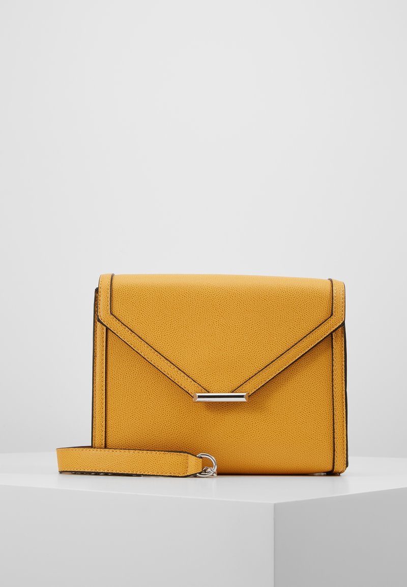 Žlutá texturovaná kožená kabelka typu clutch s obálkovým klopou, stříbrným zapínáním, černým okrajem a odnímatelným zápěstním popruhem na bílé ploše.