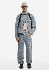 Sinivalkokuvioitu farkkutakki ja housut, vaaleanpunainen paita alla, asusteena beige lippis ja reppu.