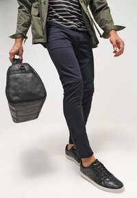 Mężczyzna w czarnych trampkach, granatowych spodniach, w pasiastą koszulę i zieloną kurtkę, trzyma czarny skórzany plecak, idąc.