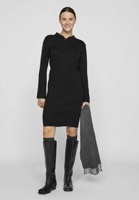 Črna obleka z dolgimi rokavi in mandarin ovratnikom, okrašena s stranskim gumbom, v kombinaciji z črnimi usnjenimi škornji do kolen in sivo šalom.