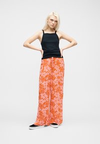 Jeune femme aux longs cheveux blonds portant un haut noir sans manches, un pantalon large à fleurs orange vif et des chaussures à enfiler noires.