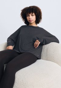 Camisa oversized com padrão escuro, decote e mangas longas, combinada com leggins pretas. Modelo sentado num sofá claro com textura.