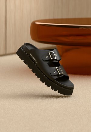 Sandalo nero con due fibbie argento, suola robusta e marchio "Karl Lagerfeld" sul lato, posizionato vicino a un tavolo rotondo marrone su pavimento beige.