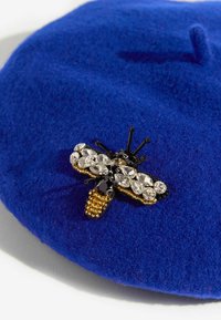 Zila filca berete, kas rotāta ar detalizētu bišu brošu, kurai ir melni un caurspīdīgi rhinestones, zelta pērlītes un melni akcenti uz spārniem.