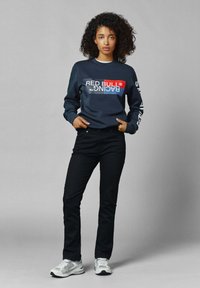 Sudadera de marina con el logo "RED BULL RACING", jeans ajustados negros y zapatillas deportivas grises con detalles de malla. Modelo de pie contra un fondo gris.