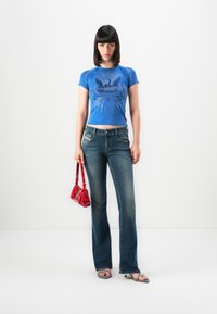 Woman with straight dark hair wearing a blue graphic t-shirt, dark flared jeans, strappy heeled sandals, holding a red patterned handbag.