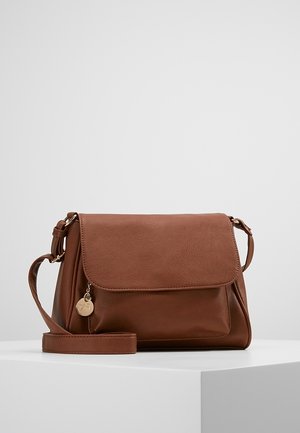 Sac bandoulière en cuir marron avec fermeture à rabat, sangle réglable et petit pendentif rond en or sur une surface blanche unie.