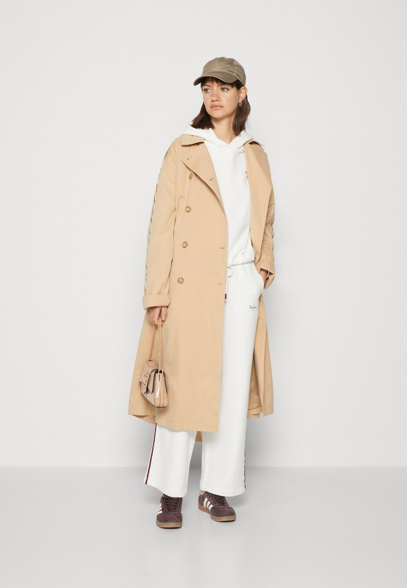 Mujer con abrigo trench beige sobre sudadera blanca y pantalones de pierna ancha, gorra beige, sosteniendo un bolso beige, con zapatillas marrones, de pie frente a una pared blanca.