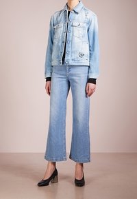 Jasnoniebieska kurtka dżinsowa z postarzanymi detalami, zestawiona z jasnoniebieskimi rozszerzanymi jeansami i czarnymi butami na obcasie. Pod spodem prosty czarny top.