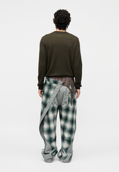 Άνδρας με σγουρά μαλλιά που φοράει σκούρο πράσινο μακρυμάνικο πουκάμισο και υπέροχο καρό παντελόνι με στρωματοποιημένη λεπτομέρεια υφάσματος, στέκεται με την πλάτη γυρισμένη σε λευκό φόντο.