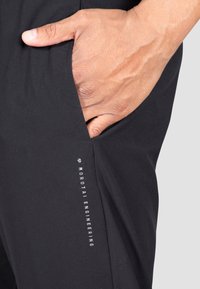 Schwarze Sporthose mit seitlicher Tasche, glatter Textur und einem vertikalen weißen Logo-Druck entlang des linken Beins.