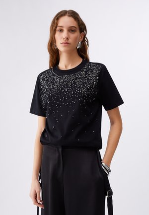 Junge Frau mit langen Haaren, die ein schwarzes kurzärmeliges Oberteil mit verstreuten silbernen Perlen und schwarze Hosen mit weitem Bein trägt.