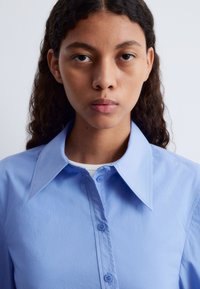 Light blue button-up shirt with a broad collar and front buttons. Smooth texture and fitted design, worn over a white undershirt.