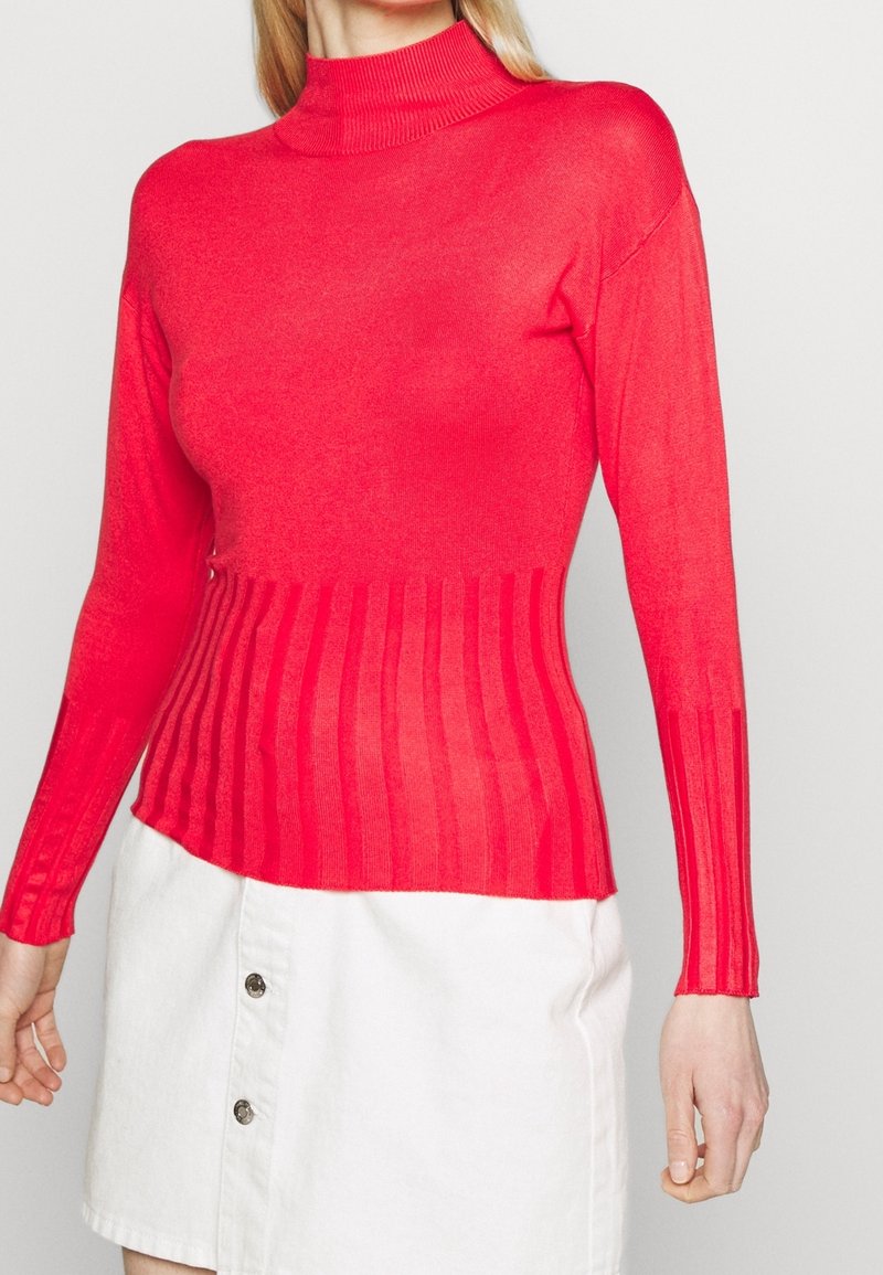 Femme portant un pull col roulé rouge ajusté à côtes verticales et une jupe blanche à boutons sur fond uni.