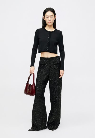 Pull cropped noir avec fermeture à bouton, associé à un pantalon large en sequins noirs. Le modèle porte un sac à main bordeaux.