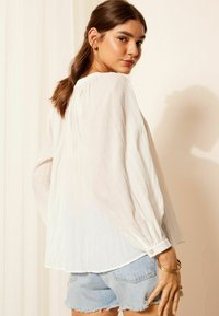 Hvid, langærmet bluse med rynket halsudskæring og struktureret stof, parret med lyseblå denimshorts. Armbåndsdetaljer på håndleddet.