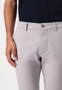 Helle graue Hose aus weichem Stoff, mit flachem Bund, Gürtelschlaufen und einem einzelnen Knopfverschluss. Enthält dezente Nähdetails.