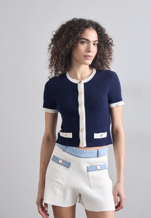 Marine gebreide cardigan met korte mouwen, afgewerkt met crème details, gouden knopen en twee grote zakken; gecombineerd met witte shorts met denimdetails.