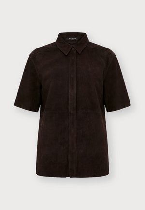 Personne aux cheveux blonds courts portant une chemise noire à manches courtes, une jupe noire longueur genou, et des bottes montantes marron foncé, debout face à l'avant.