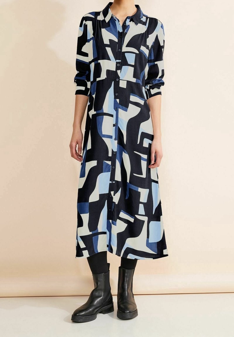 Robe à manches longues avec boutonnière sur le devant, motifs abstraits bleus, noirs et blancs, portée avec des leggings noirs et des bottines noires.
