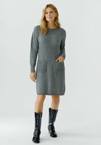 Robe en tricot grise à manches longues, deux poches avant, accents côtelés et encolure ronde, associée à des bottes noires à hauteur genou.