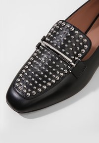 Svarta lädermockasiner med en nitar övre och metalliska kupol detaljer samt ett spänne i silverfärg. Rundad tå och slät textur.