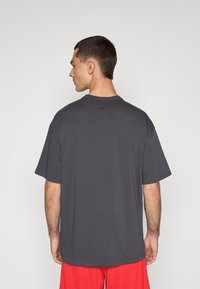 Grå kortærmet t-shirt lavet af bomuld, med rund hals og et diskret logo på den øverste ryg. Båret med røde shorts.