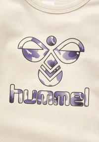 Cremefarvet stof med et lila- og blåt camouflagemønster integreret i et grafisk design. Ordet "hummel" er trykt nedenunder.
