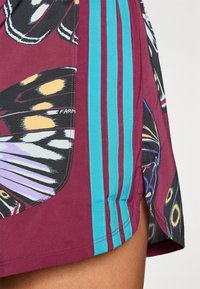 Tissu bordeaux avec imprimé d'ailes de papillon et trois rayures verticales turquoise sur un vêtement près de la cuisse d'une personne.