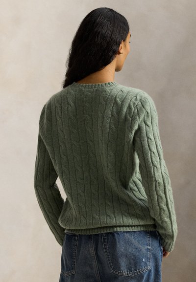 Donna con capelli lunghi e scuri che indossa un maglione verde chiaro a trecce e jeans blu, vista di spalle contro uno sfondo beige uniforme.