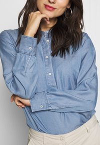 Light blue denim shirt with a button-up design, long puffed sleeves, and a relaxed fit. Two buttons visible at the cuffs.