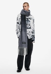 Gray knitted scarf with fringes, made of soft material, displayed with a black and white graphic shirt and dark pants. Branding visible.