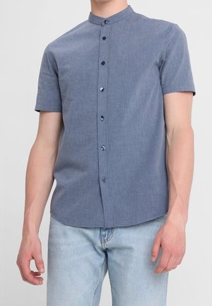 Mand iført en tætsiddende, kortærmet, blå skjorte med knapper og en bandekrave samt lyseblå jeans.