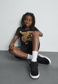 T-shirt graphique noire avec imprimé jaune, shorts noirs, chaussettes blanches montantes et baskets noires en tissu texturé avec semelle blanche. Pose assise.