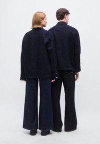 Σκούρα τζιν μπουφάν που φοριούνται από δύο μοντέλα, με άνετη εφαρμογή, ορατές ραφές, γιακά και φαρδιές παντελόνια από σκούρο τζιν με αντίθετες ραφές.