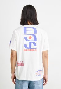 Balta trumpa rankovė t-shirt su spalvingais grafiniais spaudiniais ant nugaros, kuriuose yra mėlynas ir raudonas tekstas bei logotipai. Minkšta medvilnė.
