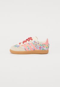 Sneaker with a cream suede upper, pink shoelaces, and colorful floral embroidery. Features three pink stripes and a brown rubber sole.
