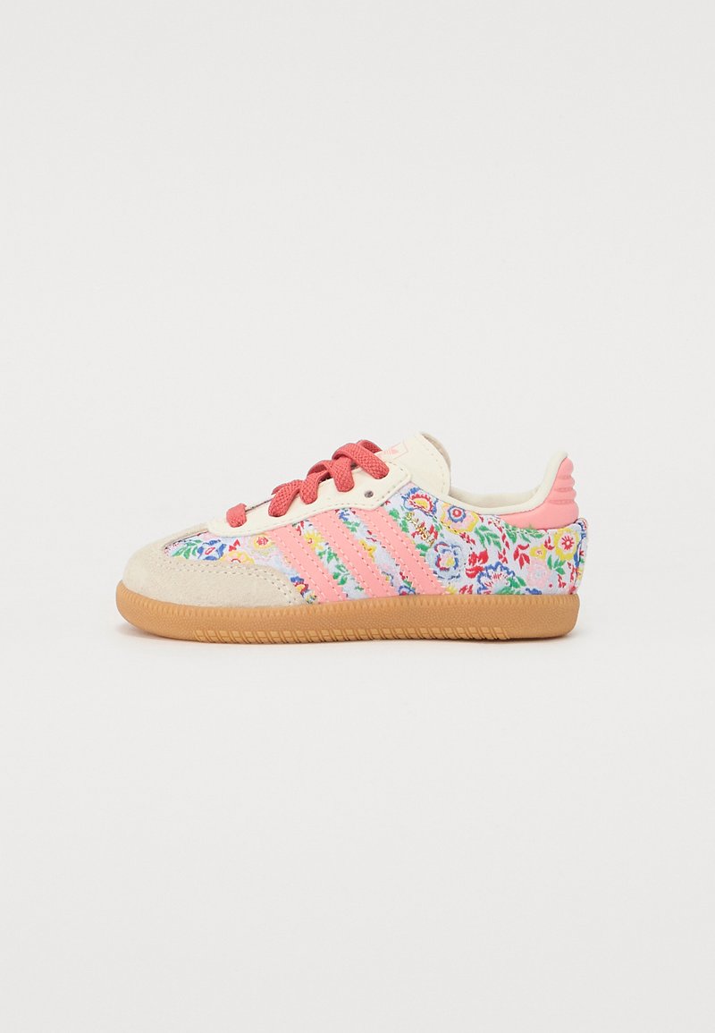 Sneaker with a cream suede upper, pink shoelaces, and colorful floral embroidery. Features three pink stripes and a brown rubber sole.