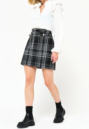 Kobieta ubrana w białą bluzkę z falbankami, czarno-białą spódnicę w kratę z guzikami, czarne skarpetki i masywne czarne botki.