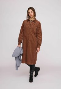 Rochie cămașă maro din catifea cu nasturi în față, mâneci lungi și buzunar la piept, asortată cu un pulover tricotat gri deschis. Cizme negre până la genunchi.