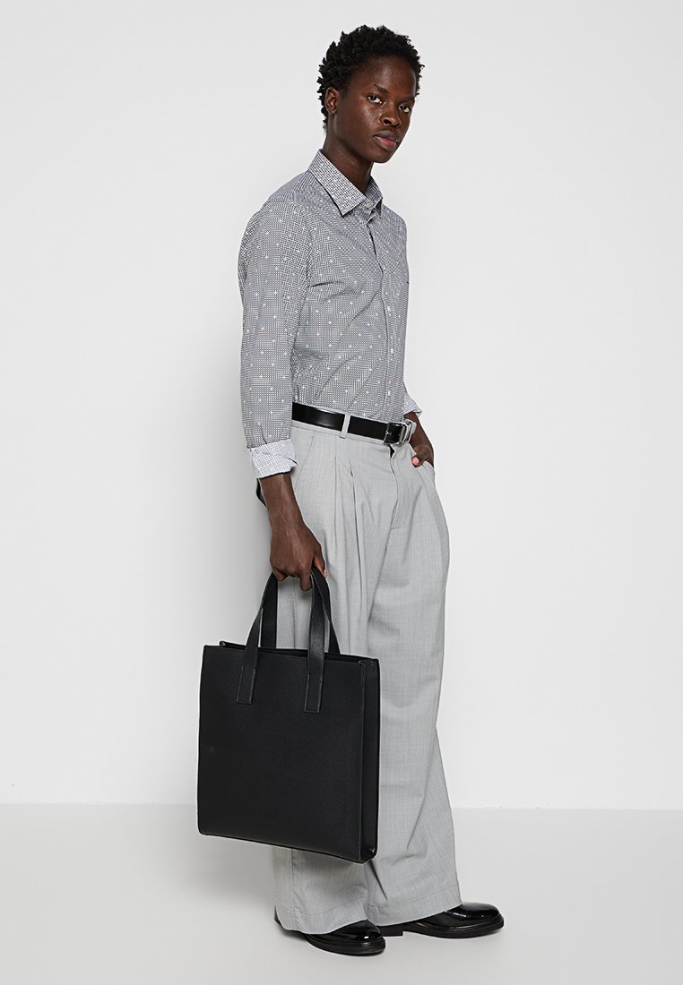 Bolso tote de cuero negro con dos asas, textura suave, combinado con una camisa gris con estampado y pantalones plisados gris claro.