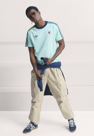 Jeune homme portant un t-shirt Adidas bleu clair, un pantalon cargo beige, un sweat-shirt bleu marine attaché à la taille et des baskets bleues, debout devant un fond uni.