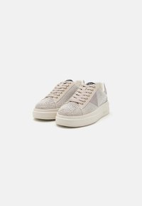 Sneaker mit einem beigefarbenen Stoff-Obermaterial, verziert mit silbernen Nieten, und einer dicken weißen Sohle sowie tonalen Akzenten. Schnürsenkel und Logodetail sind enthalten.