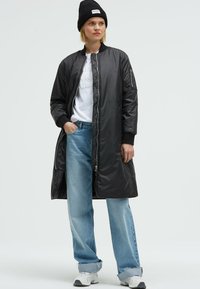 Black padded long coat with a zip front, ribbed cuffs, and side pockets, worn over a white T-shirt and light blue wide-leg jeans.