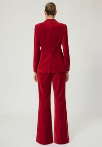 Rød fløjlssuite med en tætsiddende blazer og skrædderbukser. Blazeren har en klippet krave og knapdetaljer. Glat tekstur hele vejen igennem.