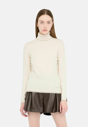 Cremfarbener Rollkragenpullover mit geripptem Halsausschnitt und Bündchen, kombiniert mit dunkelbraunen plissierten Shorts aus Kunstleder. Markenlogo am Saum des Pullovers.