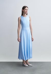 Junge Frau steht, trägt ein ärmelloses, hellblaues Midikleid und weiße spitze Absatzschuhe, vor einem schlichten grauen Hintergrund.