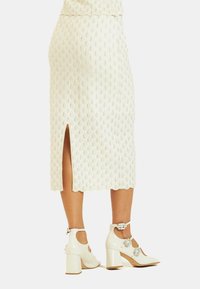 Weißer Strick-Midi-Rock mit strukturiertem Rautenmuster und seitlichem Schlitz. Kombiniert mit weißen Lackblockabsatzschuhen mit dekorativen Akzenten.