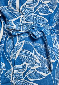 Tessuto blu con una finitura testurizzata, caratterizzato da un motivo foglioso bianco. Include un dettaglio con cintura annodata in vita.